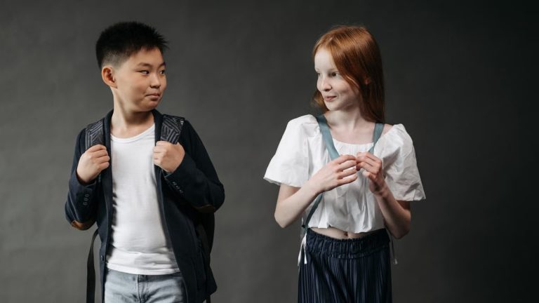 Two children wearing backpacks look at each other, standing indoors with a neutral background.